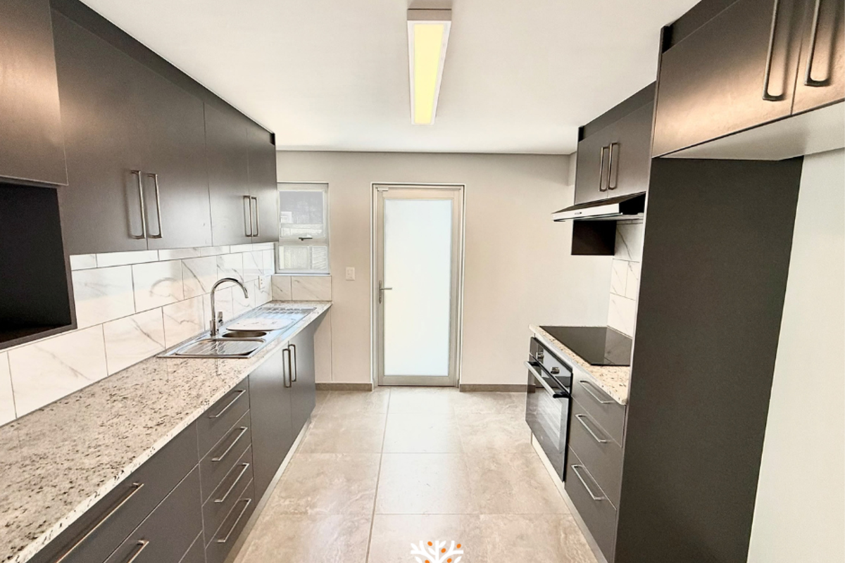 Modern galley-style kitchen featuring dark cabinetry, granite countertops, built-in oven, electric hob, and sleek tiled flooring with direct access to the exterior.