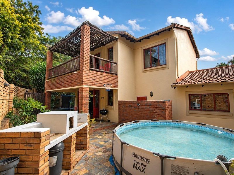 Rear exterior view of the townhouse featuring a brick built-in braai, tiled patio area, and an upper-level balcony with a timber pergola.