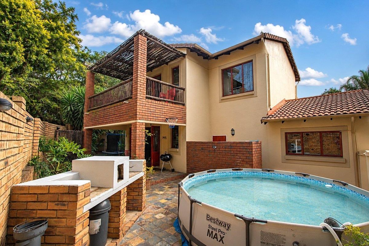 Rear exterior view of the townhouse featuring a brick built-in braai, tiled patio area, and an upper-level balcony with a timber pergola.
