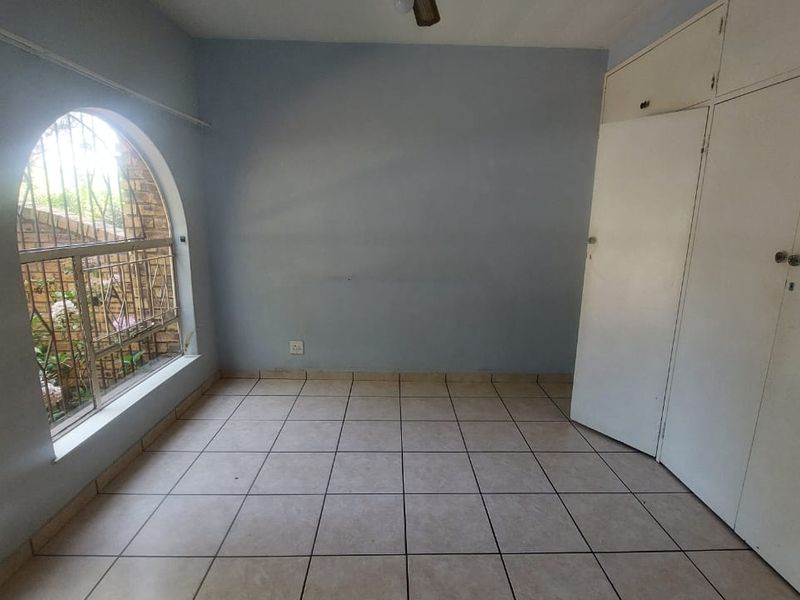 An empty bedroom featuring tiled flooring, built-in cupboards, and a distinctive arched window with security bars.