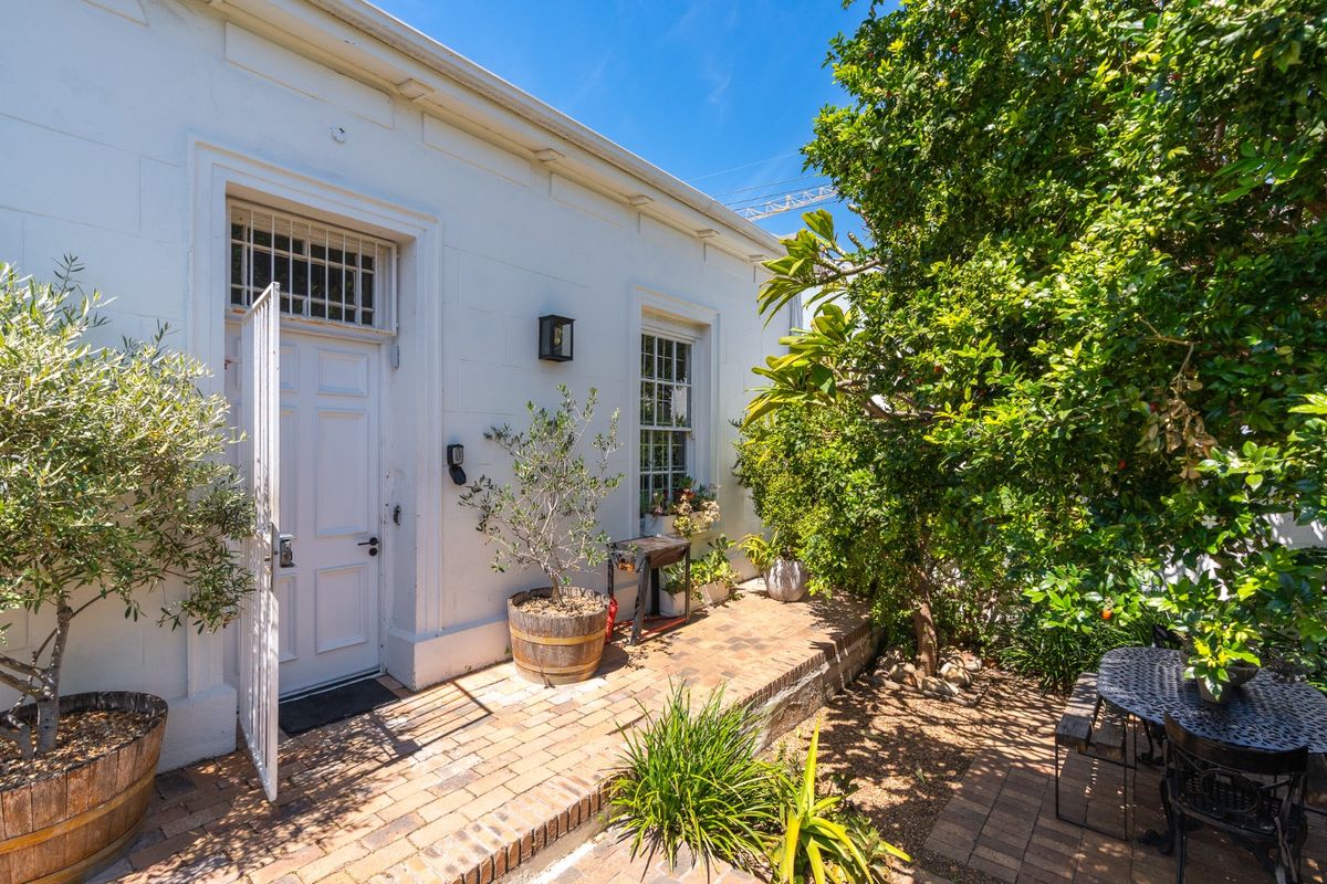 A charming rear exterior view featuring a brick-paved patio, white painted facade, and classic sash windows, surrounded by established garden greenery.