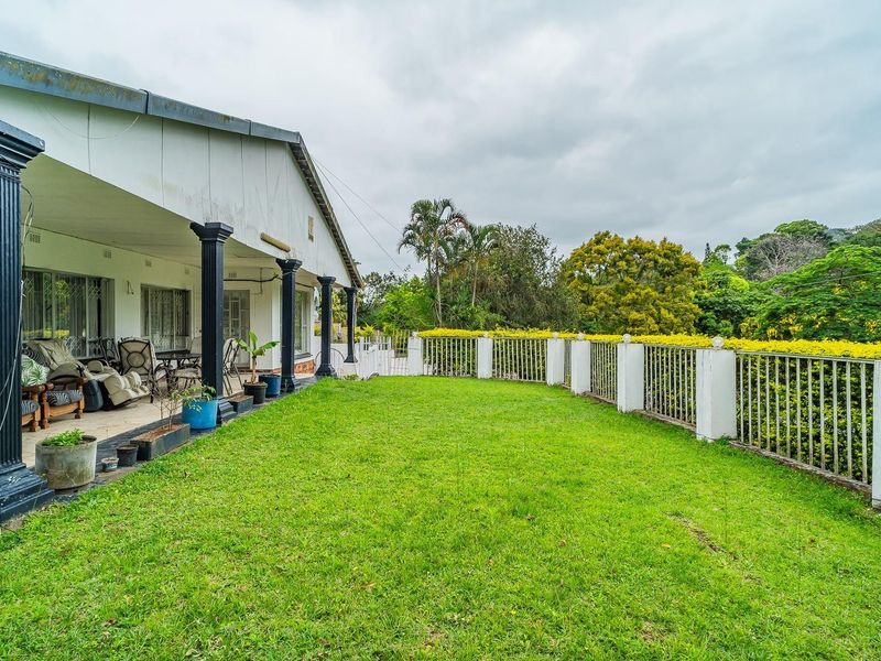 Rear view of the property featuring a covered patio with decorative columns, overlooking a spacious lawn and secure perimeter fencing.