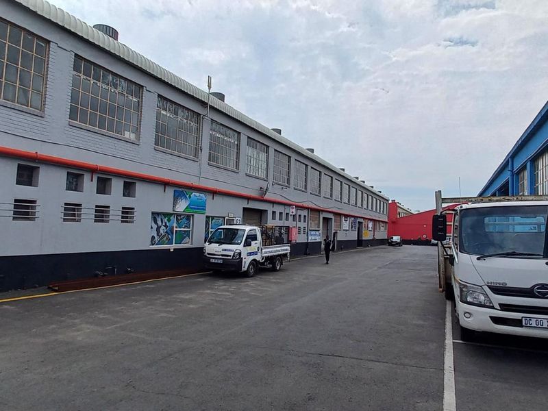 Exterior view of an industrial warehouse facility featuring a large asphalt yard, steel-framed windows, and exposed fire safety infrastructure.