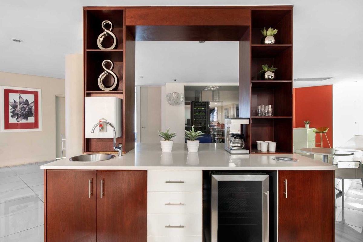 A modern office kitchenette featuring dark wood cabinetry, integrated wine cooler, stone countertop, and a built-in sink with a chrome faucet.