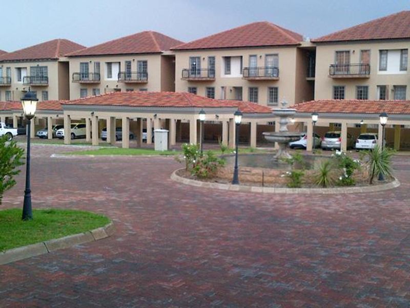 Exterior view of a townhouse complex featuring a paved driveway, covered parking bays, and a central decorative water fountain.