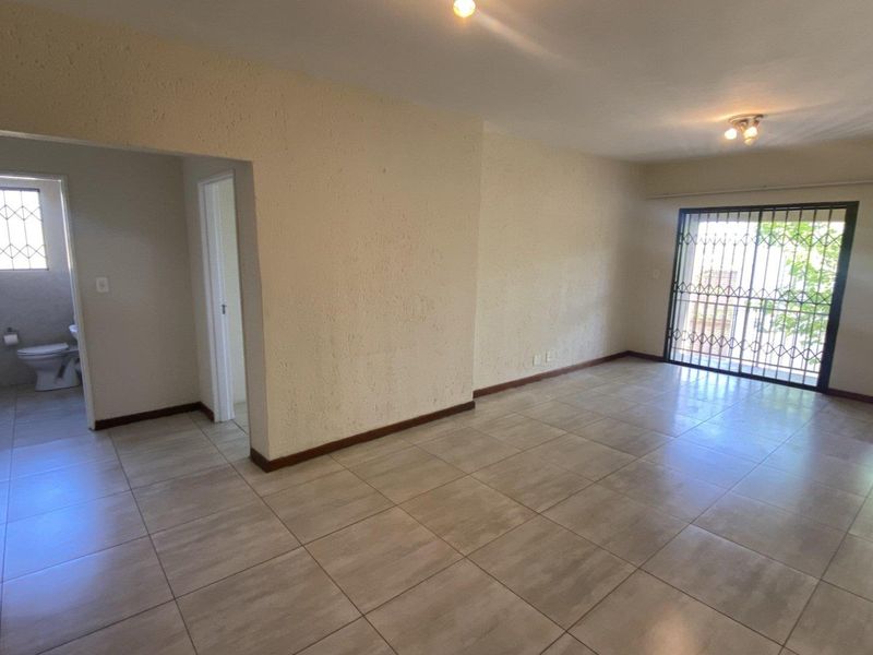An empty, open-plan living area featuring neutral tiled flooring, security gates on windows, and direct access to an adjacent bathroom.