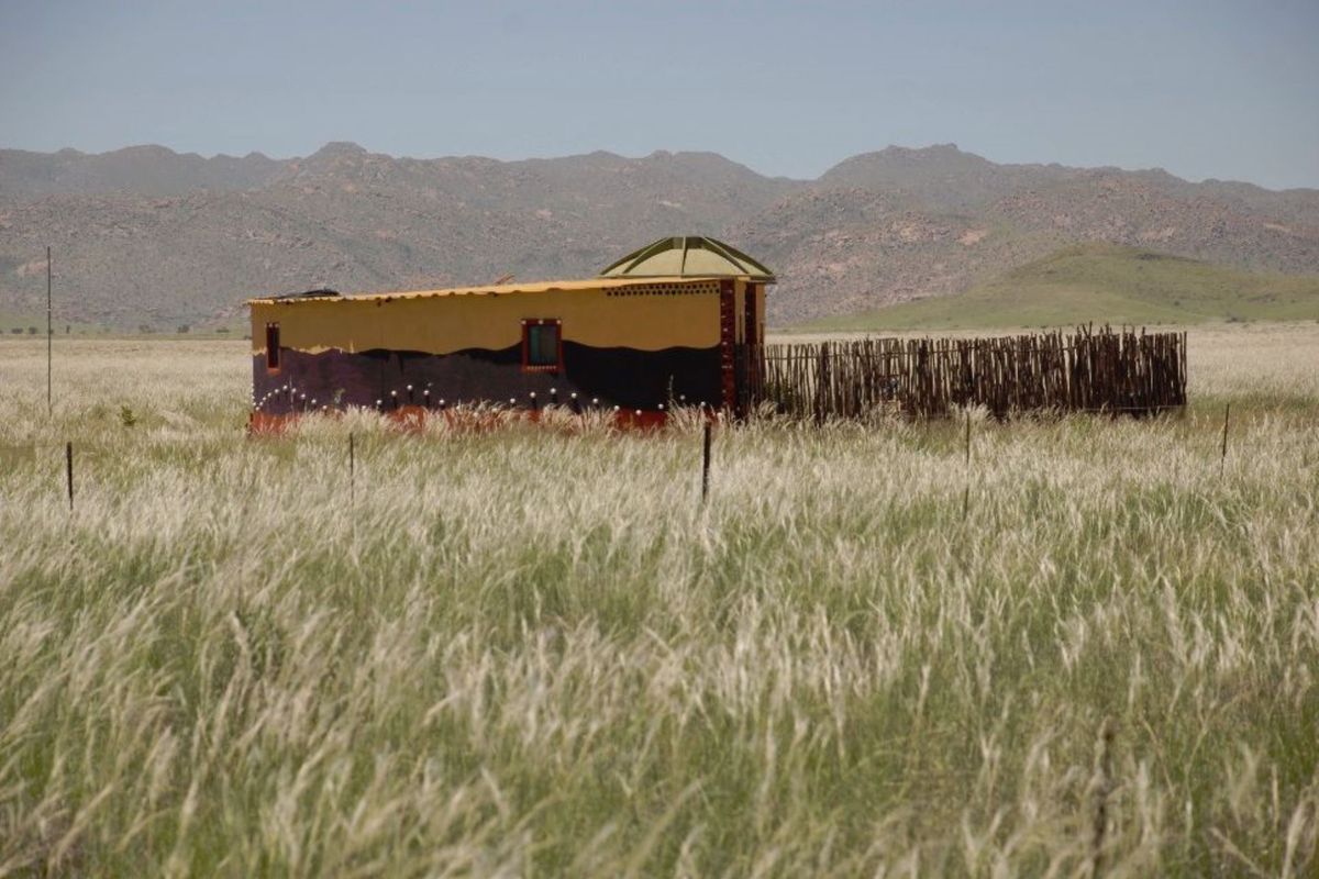 A small, uniquely painted rural structure set within a vast grassy field, featuring a distinctive domed roof section and rustic perimeter fencing.