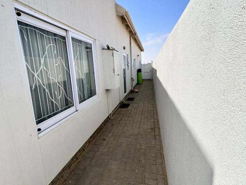 A narrow paved side passage along the exterior of the house, featuring a boundary wall and sliding windows with security bars.