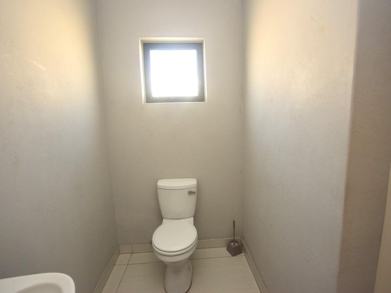 A compact, minimalist guest bathroom featuring tiled flooring, a standard toilet, and a small window for natural light.
