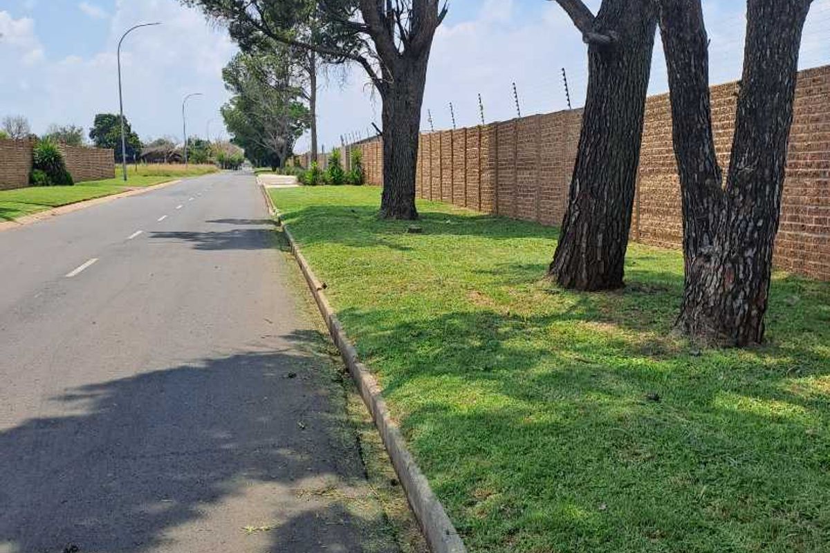 A view of the street frontage featuring a well-maintained brick boundary wall with electric fencing, mature trees, and a paved road.