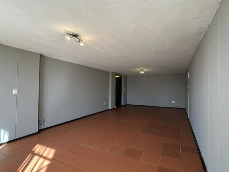 A spacious, empty living area featuring neutral grey walls and terracotta-style tiled flooring, illuminated by ceiling-mounted light fixtures.