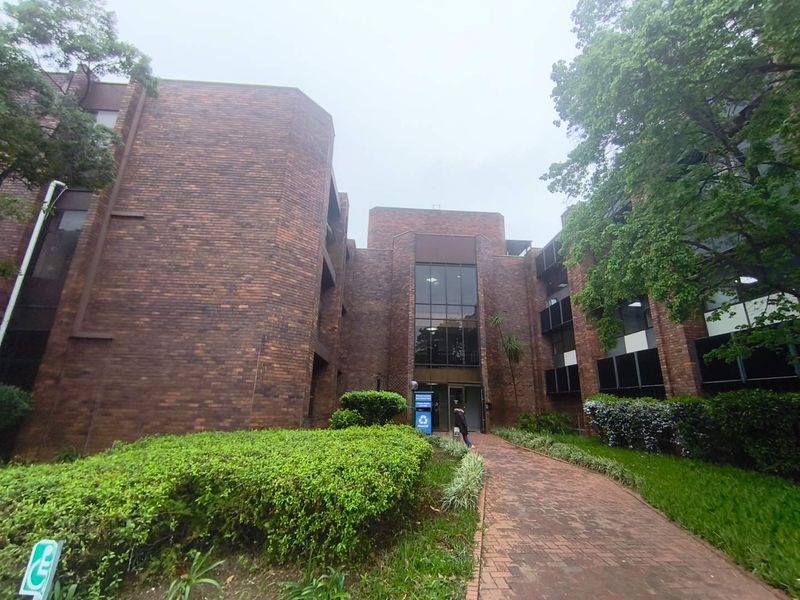 Front exterior view of a multi-story brick office building featuring a paved walkway, landscaped garden, and a prominent glass entrance.