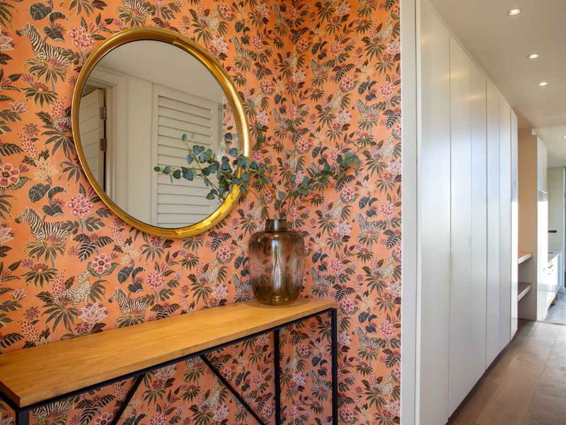 A well-lit hallway featuring floor-to-ceiling built-in cabinetry, warm timber flooring, and decorative wallpaper, leading towards an open-plan living area.
