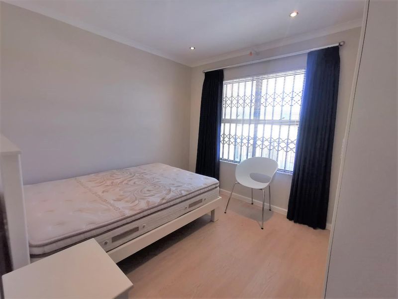 A well-lit bedroom featuring light laminate flooring, recessed downlighting, and a window fitted with security bars and dark curtains.