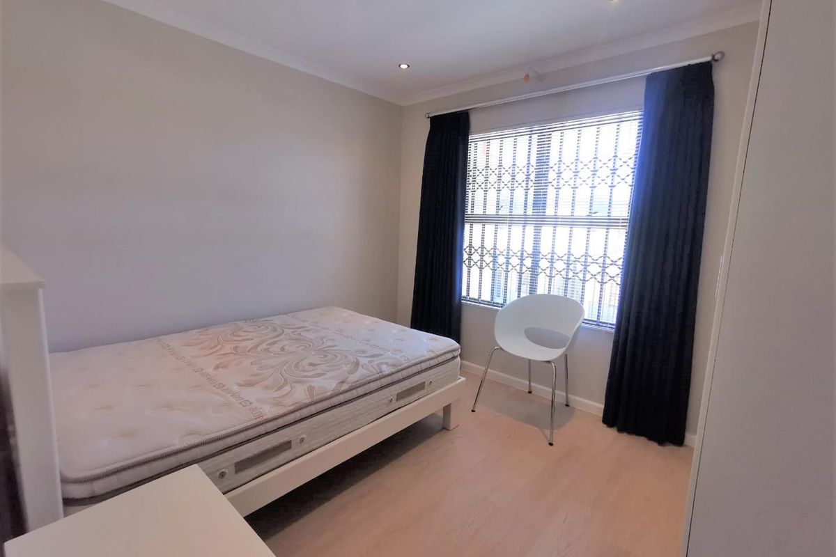 A well-lit bedroom featuring light laminate flooring, recessed downlighting, and a window fitted with security bars and dark curtains.