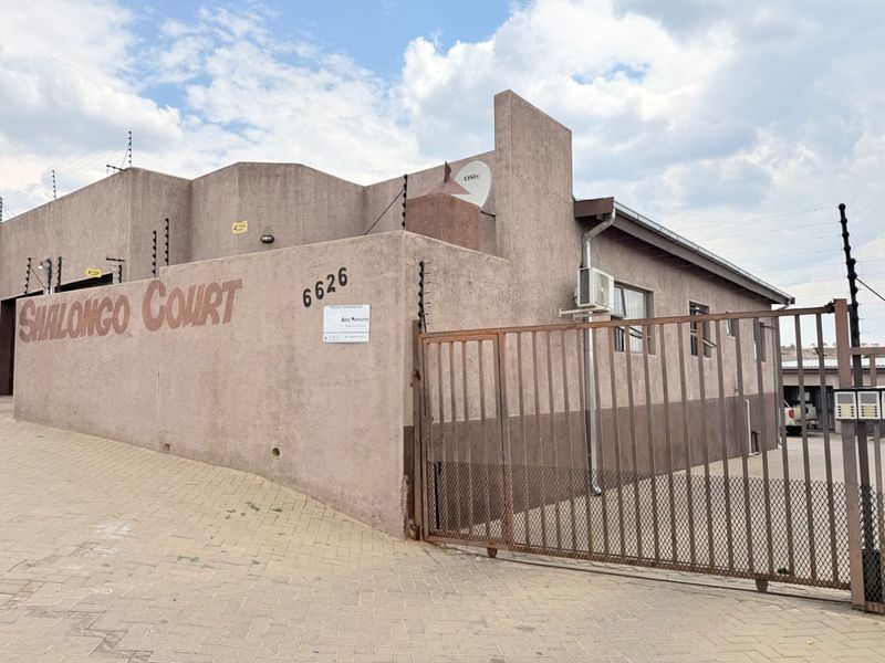 Exterior view of Shalongo Court townhouse complex featuring a paved driveway, secure metal sliding gate, and perimeter electric fencing.