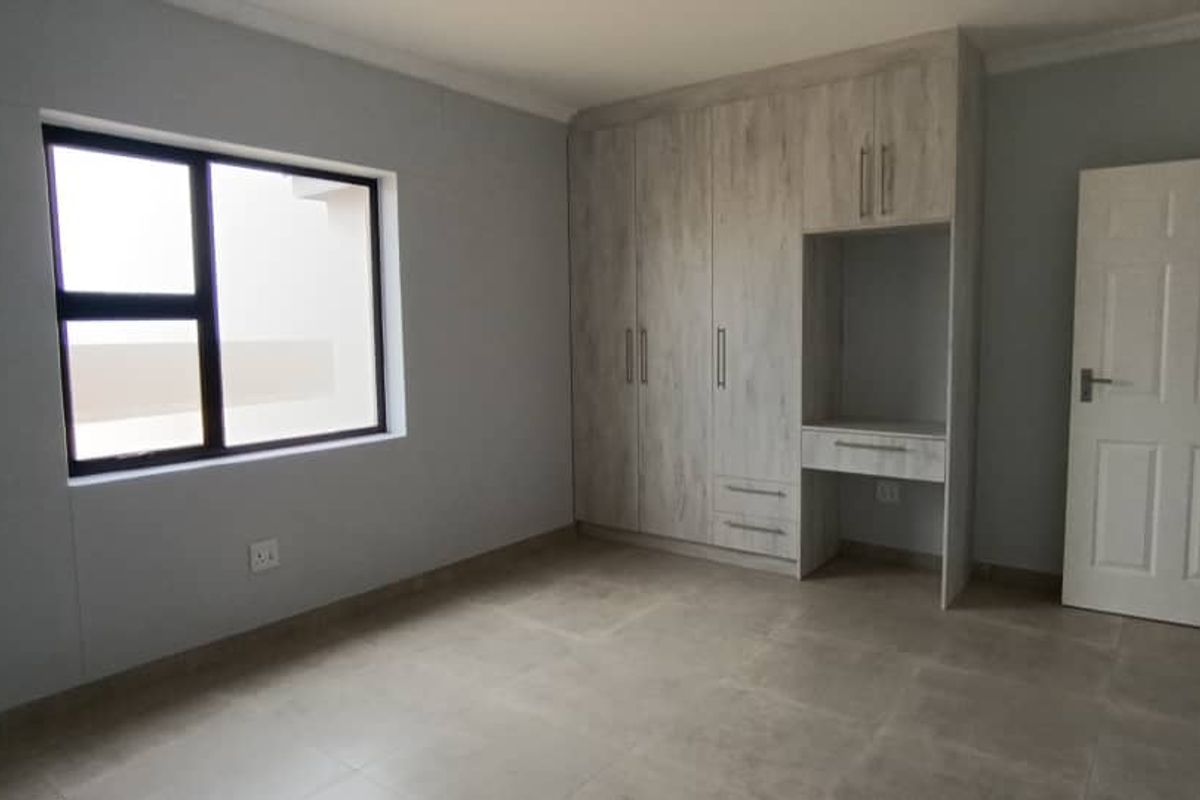 A bright, modern bedroom featuring neutral floor tiles, built-in wooden cabinetry with an integrated desk, and a large window providing natural light.