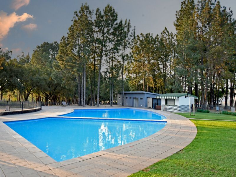 A well-maintained outdoor swimming pool area featuring a paved deck, surrounding lawn, and a small kiosk structure set against a backdrop of mature trees.
