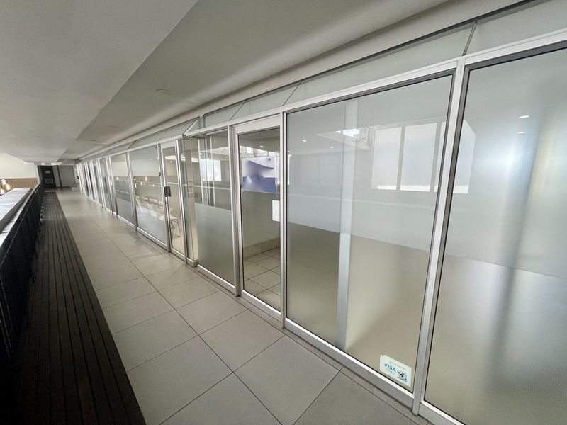 A bright, modern commercial hallway featuring extensive glass partitioning, aluminum framing, and clean tiled flooring, suitable for office or retail use.
