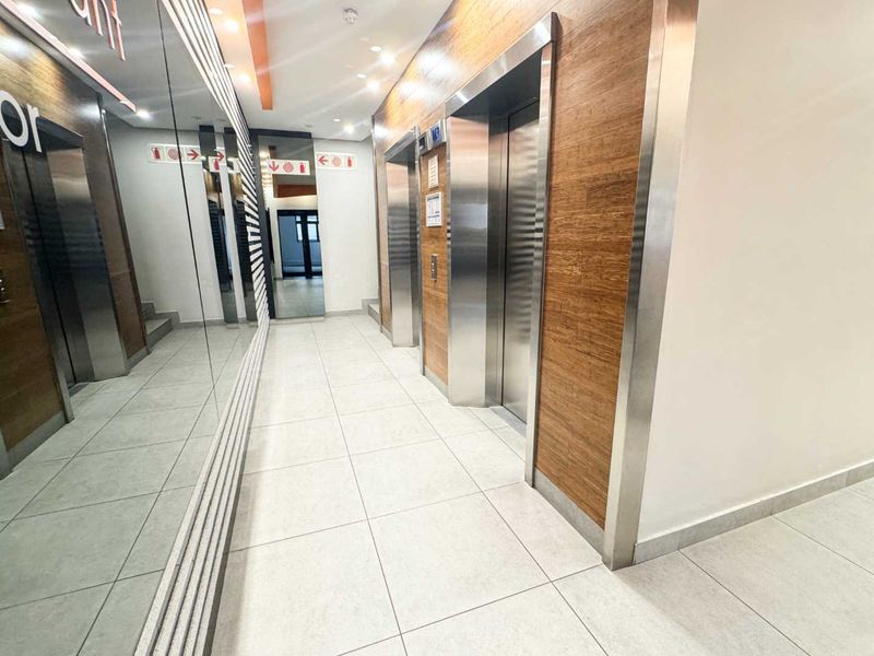 Modern office building elevator lobby featuring stainless steel lift doors, wood-paneled walls, large mirrors, and clean tiled flooring.