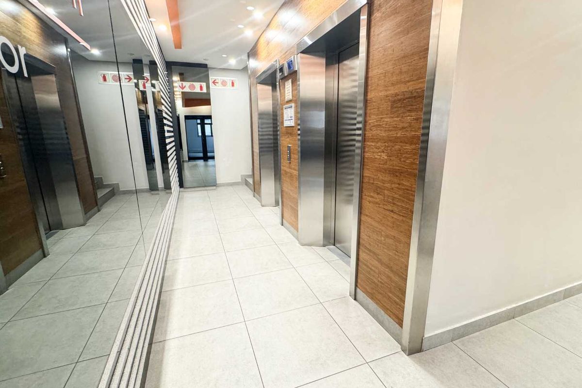 Modern office building elevator lobby featuring stainless steel lift doors, wood-paneled walls, large mirrors, and clean tiled flooring.