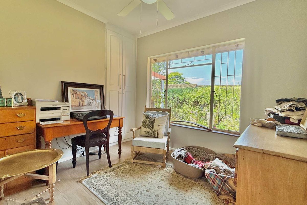 A bedroom featuring built-in cupboards, a ceiling fan, and a large window with security bars overlooking the garden.