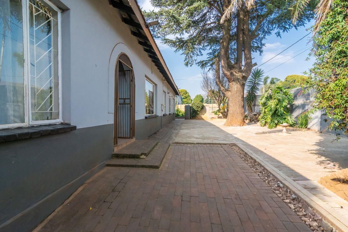 A view of the rear exterior of the house featuring a paved patio area, mature trees, and a secure arched doorway.