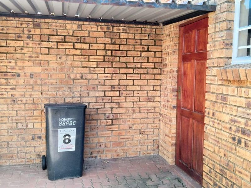 A covered exterior entrance area featuring face brick walls, a timber door, and paved flooring, providing a functional access point to the townhouse.