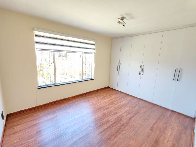 A bright, empty bedroom featuring laminate flooring, a large window with a roller blind, and extensive built-in cupboard storage.