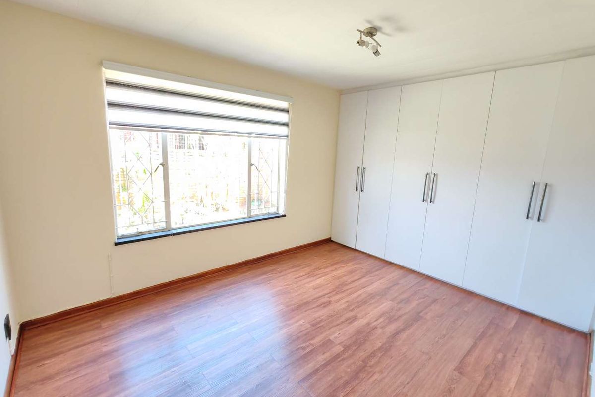 A bright, empty bedroom featuring laminate flooring, a large window with a roller blind, and extensive built-in cupboard storage.