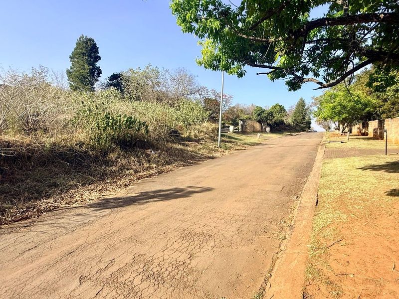 A view of the residential street approach featuring an asphalt road, mature trees, and established boundary walls.