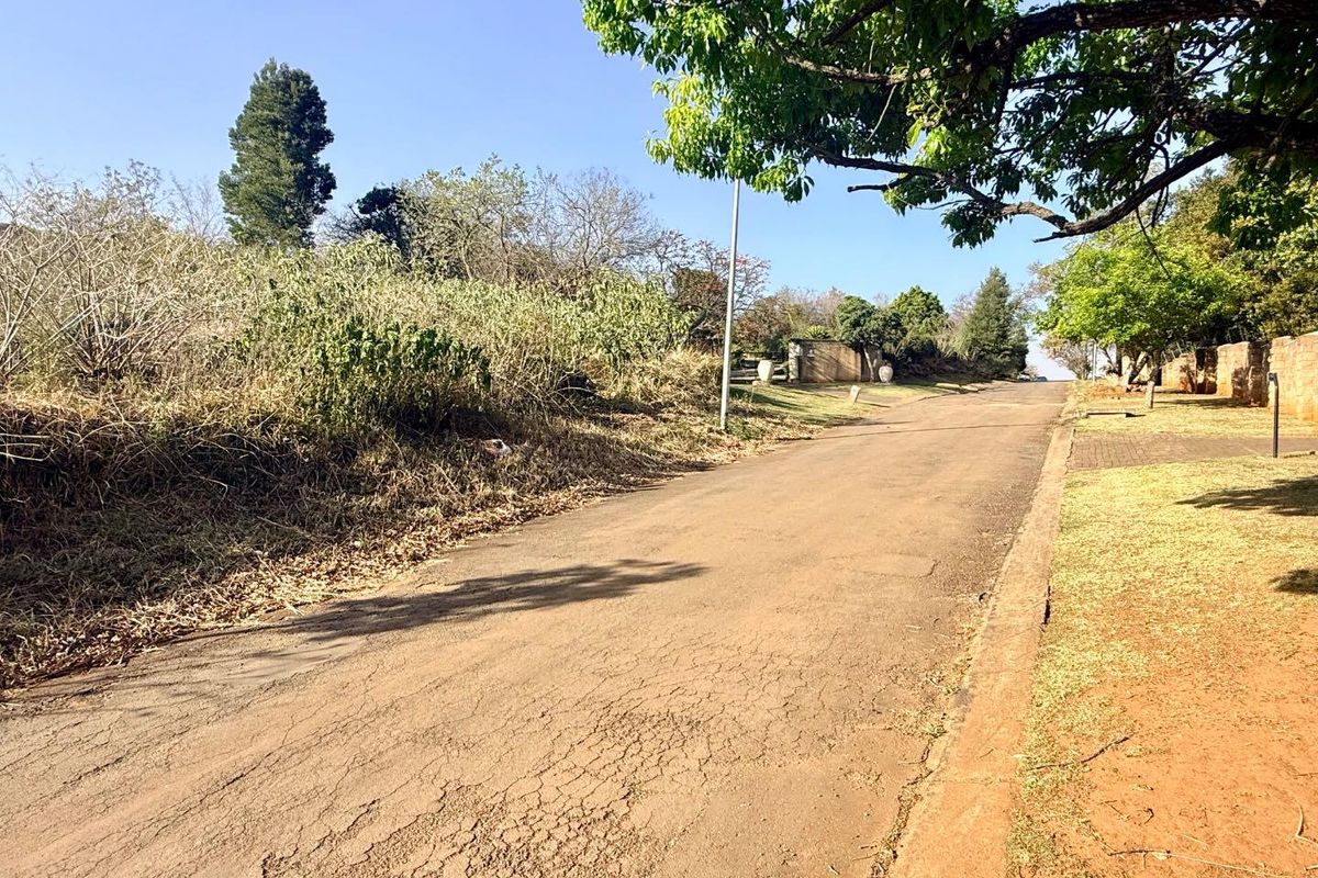 A view of the residential street approach featuring an asphalt road, mature trees, and established boundary walls.