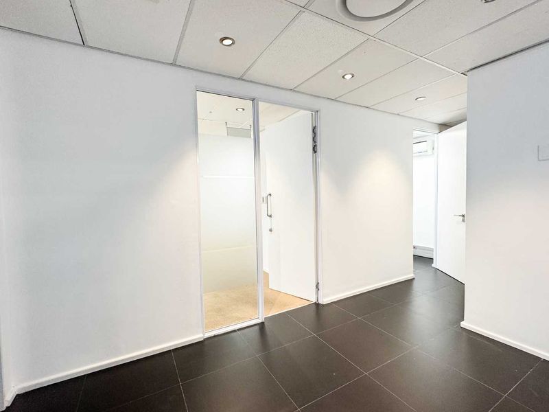 A clean, modern office hallway featuring dark tiled flooring, a suspended ceiling with recessed lighting, and a glass partition door.