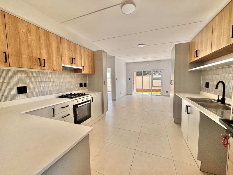Modern galley-style kitchen featuring light wood cabinetry, stone countertops, gas hob, electric oven, and tiled flooring, leading into an open-plan living area.