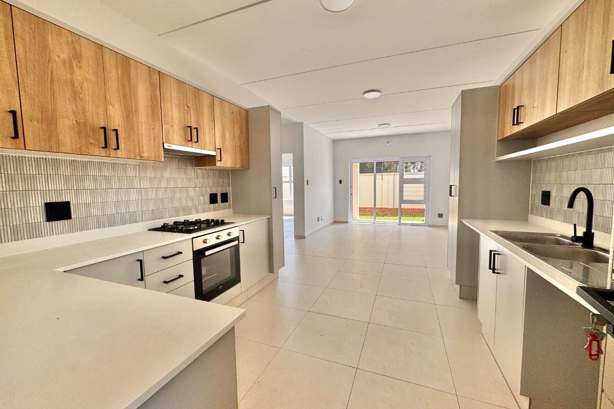 Modern galley-style kitchen featuring light wood cabinetry, stone countertops, gas hob, electric oven, and tiled flooring, leading into an open-plan living area.