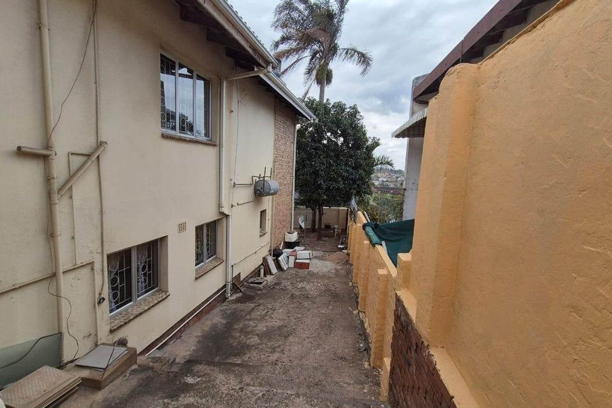 A side exterior view of the property showing a concrete driveway, boundary wall, and external geyser unit along the building facade.