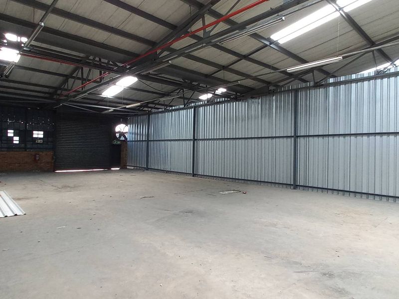 Spacious industrial warehouse unit featuring high ceilings with exposed steel trusses, corrugated metal walls, concrete flooring, and a large roller shutter door.