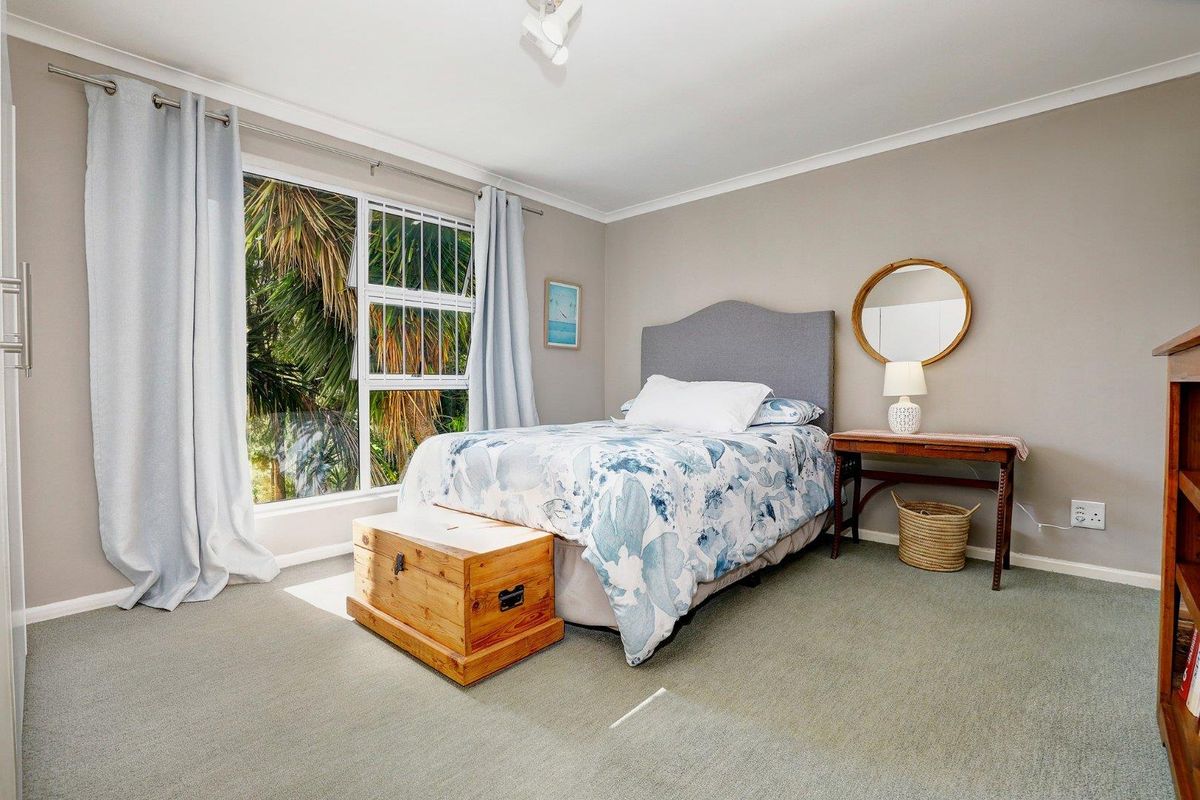 A well-lit bedroom featuring neutral wall tones, carpeted flooring, and a large window with security bars overlooking lush greenery.