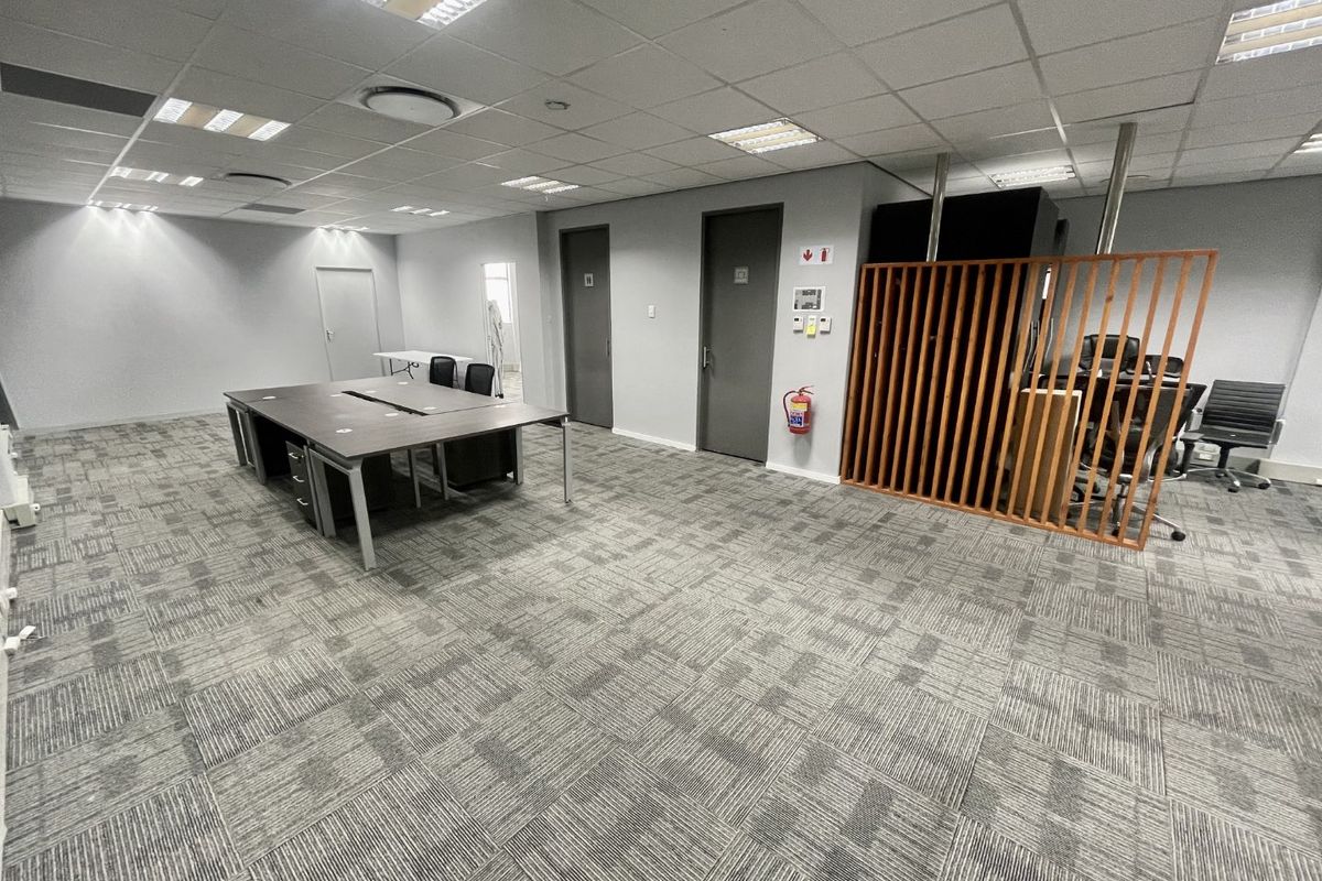 A spacious open-plan office area featuring carpeted flooring, a suspended ceiling with recessed lighting, and a decorative wooden slat partition.