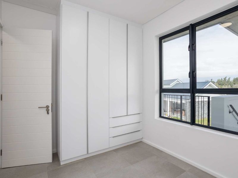A bright, modern bedroom featuring neutral tiled flooring, built-in floor-to-ceiling cupboards, and large windows with black aluminium frames.