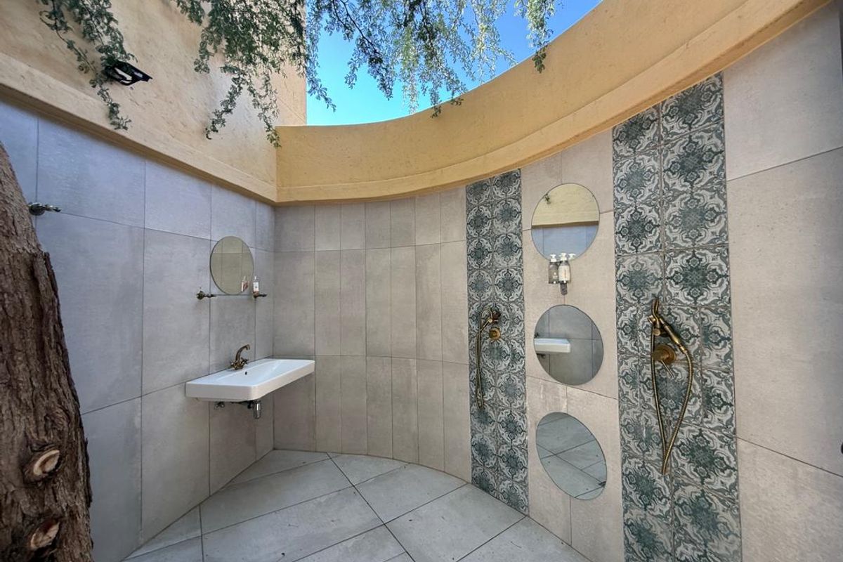 An open-air outdoor shower area featuring decorative patterned tiling, a wall-mounted basin, and dual shower fixtures with a curved architectural wall design.