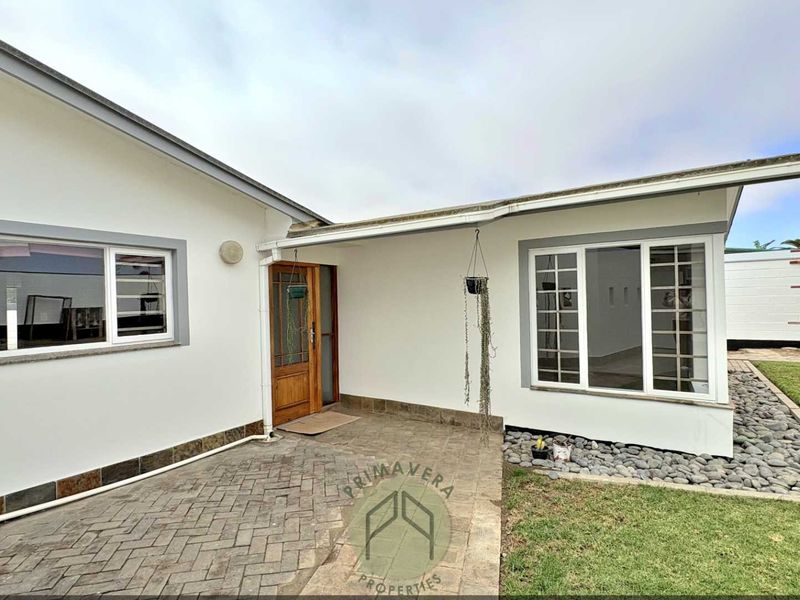A well-maintained rear exterior view featuring a paved patio area, timber entrance door, and clean white facade with modern aluminium window frames.