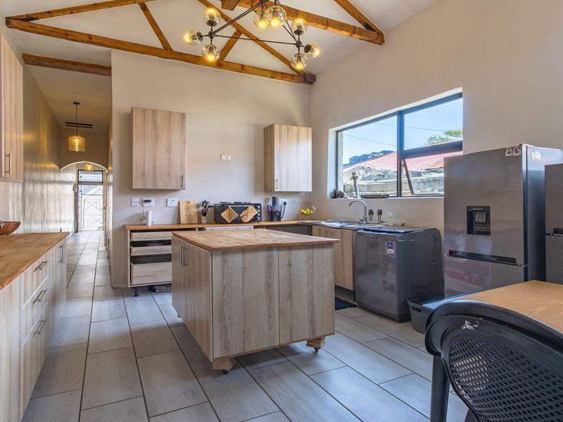 Modern kitchen featuring exposed timber roof trusses, light wood cabinetry, wooden countertops, a central island, and tiled flooring with a view into the hallway.