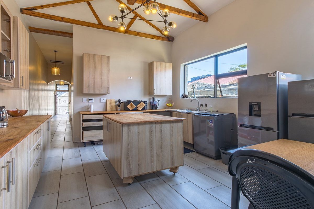 Modern kitchen featuring exposed timber roof trusses, light wood cabinetry, wooden countertops, a central island, and tiled flooring with a view into the hallway.