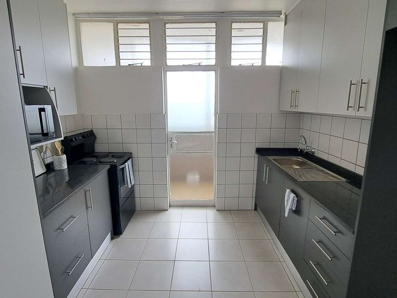 A functional galley-style kitchen featuring dark cabinetry, stone countertops, tiled backsplash, and direct access to an outdoor area through a glass-paneled door.
