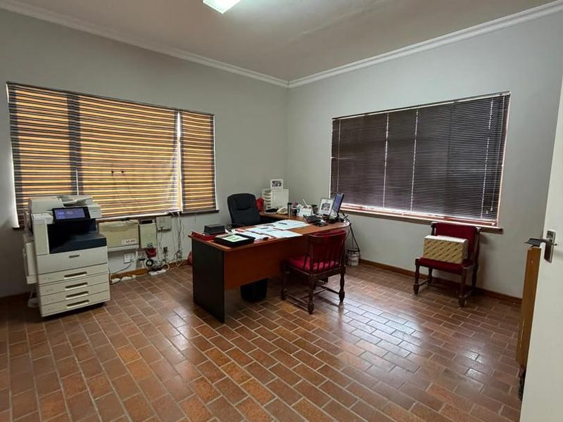 A functional office space featuring terracotta tile flooring, large windows with venetian blinds, and overhead fluorescent lighting.