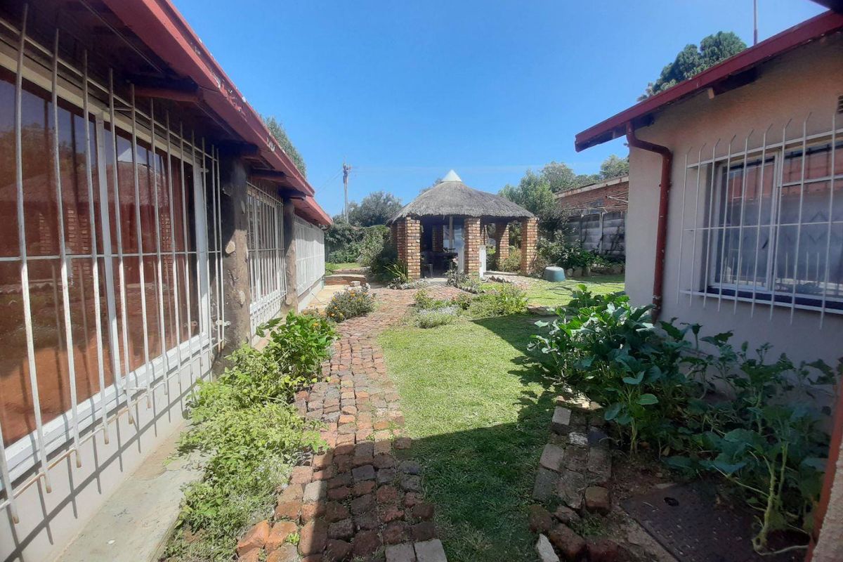 A rear garden area featuring a brick pathway, lawn, and a thatched lapa structure situated between two sections of the house.
