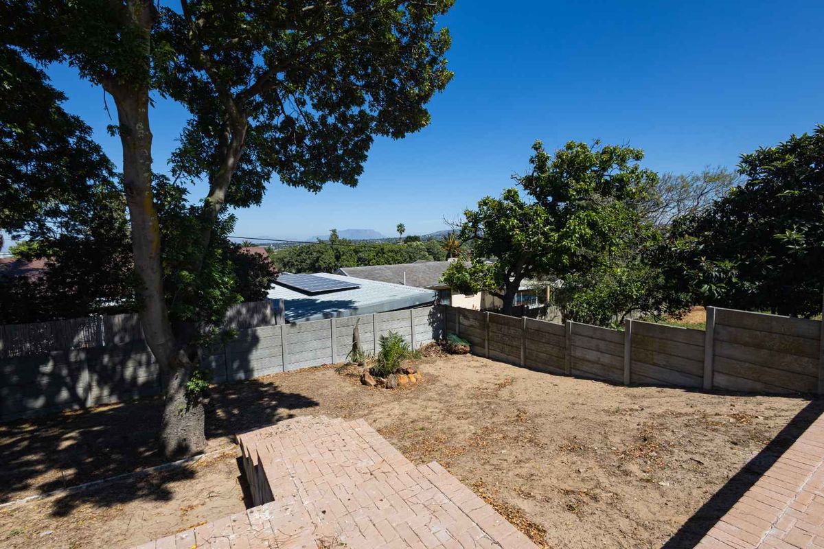 A spacious rear garden featuring a brick-paved patio area, mature trees, and a precast concrete boundary wall with views of the surrounding neighborhood.