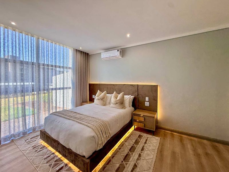 Modern bedroom featuring floor-to-ceiling windows, integrated headboard lighting, and a wall-mounted split-system air conditioner with high-quality laminate flooring.