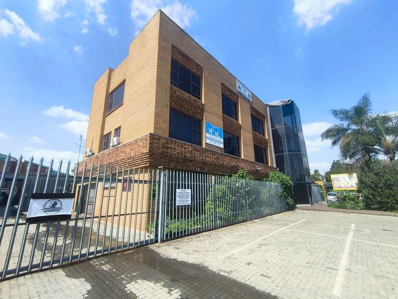 A multi-story commercial office building featuring a brick facade, glass elevator shaft, and secure gated parking area.